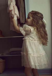 Robe rose clair avec des manches puff transparentes, des motifs brodés et une bordure en dentelle, superposée et fluide avec une texture douce. Enfant tendant la main vers des vêtements.