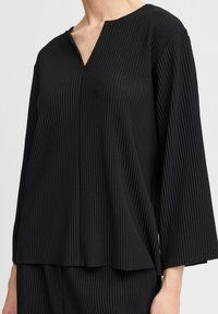 Black pleated top with a V-neckline and wide sleeves. Made of textured fabric, featuring vertical stripes and an asymmetrical hem.