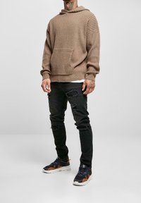 Brun ribbad hoodie med en frampå ficka, kombinerad med svarta slitna jeans och svarta sneakers med orangea och blå accentfärger.