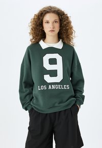 Zielona bluza z białym kołnierzykiem, z dużą białą cyfrą "9" i napisem "LOS ANGELES" w pogrubionym tekście. Miękka tekstura, luźny krój.