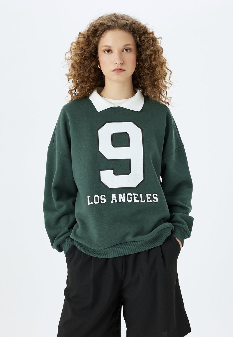 Zielona bluza z białym kołnierzykiem, z dużą białą cyfrą "9" i napisem "LOS ANGELES" w pogrubionym tekście. Miękka tekstura, luźny krój.