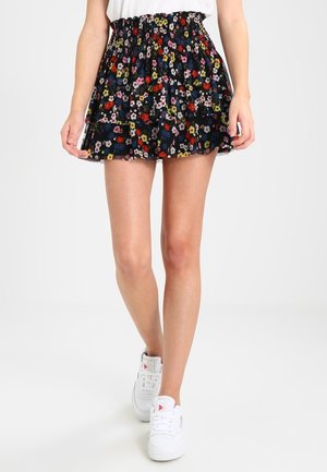 Kvinde iført en sort, blomsterprintet, høj talje mininederdel kombineret med hvide sneakers, stående mod en ensfarvet baggrund.