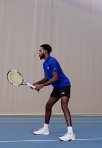 Tennis spiller i blå skjorte og svarte shorts, holder en racket. Hvite sneakers. Innendørs bane med blå overflate og beige bakgrunn.