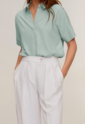 Femme portant une chemise boutonnière à manches courtes vert clair, rentrée dans un pantalon blanc taille haute, les mains dans les poches.