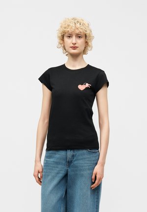 Fiatal nő göndör szőke hajjal, fekete, testhezálló pólót visel, amelyen rózsaszín szív és felirat látható, valamint kék farmert, egy egyszerű háttér előtt áll.