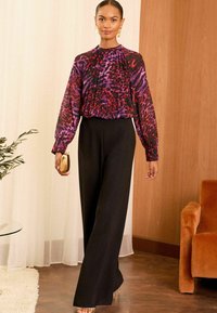 Blusa multicolore con motivo astratto viola e rosso, dettagli plissettati, abbinata a pantaloni larghi neri, mentre tiene una pochette dorata.
