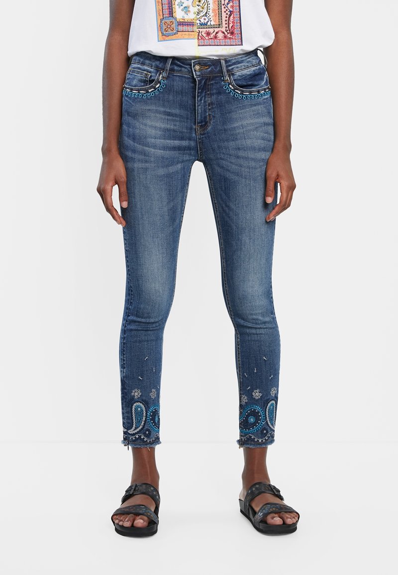 Femme portant un jean skinny bleu avec une broderie bleue et blanche près des poches et de l'ourlet, associée à des sandales noires.