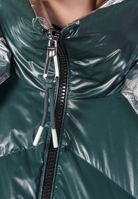 Giacca puffer verde in materiale lucido e trapuntato, con accenti argentati sulle spalle e una zip prominente con cordino e fermaglio.