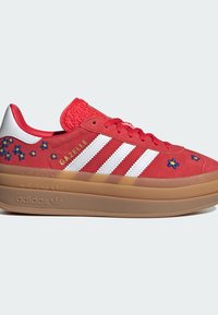 Baskets Adidas Gazelle rouges avec des bandes blanches, des fleurs bleues et jaunes brodées, et une semelle épaisse marron sur un fond clair uni.