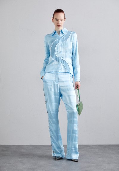 Costum din satin de culoare bleu deschis format din cămașă și pantaloni, prezentând un detaliu cu cusături ondulate și butoane decorative pe laturile pantalonilor; completat cu o geantă verde.
