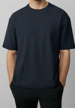 Man wearing a dark blue textured short-sleeve shirt with hands in black pants pockets against a light gray background.