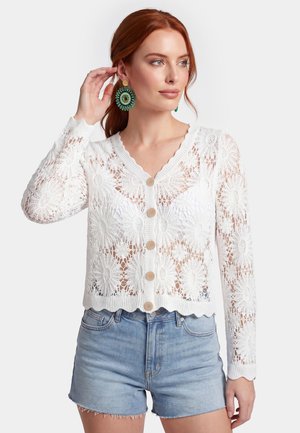Kvinde med rødt hår iført store grønne runde øreringe, en hvid blomsterhæklet knap-cardigan og lyseblå denimshorts.