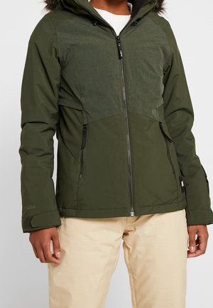 Osoba ubrana w ciemnozieloną kurtkę zapinaną na zamek z zapinanymi kieszeniami oraz beżowe spodnie, stojąca na jednolitym tle.