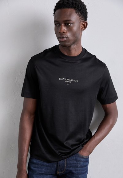 T-shirt noir à manches courtes en tissu lisse, avec un logo de marque brodé en blanc sur la poitrine, associé à un jean en denim bleu foncé.