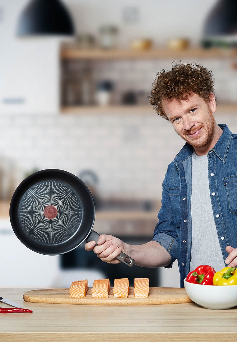Eiserne schwarze Bratpfanne mit einem roten Kernmuster, über Lachsfilets auf einem Holzschneidebrett gehalten, mit einer Schüssel Paprika in der Nähe.