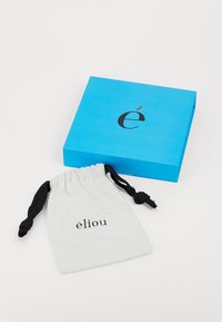 Caja azul con logo negro y acento rojo junto a una bolsa de cuerda blanca que presenta el nombre de la marca "élio" en negro.