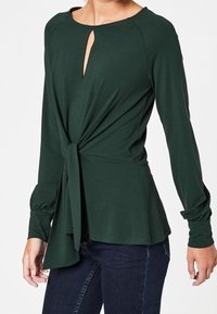 Tmavě zelený top s dlouhým rukávem, výstřihem ve tvaru V s klíčovým otvorem, detaily uzlu vpředu a nařasenými rukávy. Měkká, elastická látka.