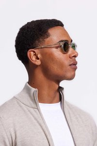 Ronde zonnebril met een heldere rand, groene lenzen en metalen accenten; model draagt een beige ritsvest over een wit shirt.