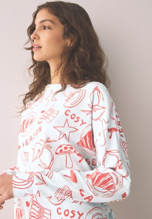 Sieviete ar gariem cirtainiem matiem valkā baltu džemperi ar sarkaniem sēņu, augļu, zvaigžņu zīmējumiem un vārdiem “COSY” un “SEASON” uz vienkārša fona sienas.