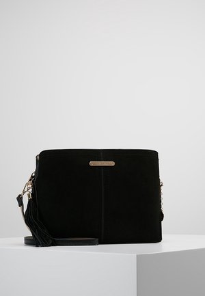 Zwarte rechthoekige crossbodytas met gouden ketting en tassel-detail op een witte ondergrond en lichte grijze achtergrond.