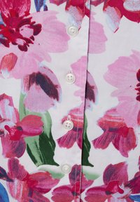 Bloes met bloemenmotief die grote roze en rode bloemen op een witte achtergrond toont. De knopen zijn wit en rond, met een gladde stofstructuur.