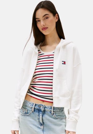 Hoodie bianca croppata con zip completa, dotata di tasca frontale e polsini a coste, abbinata a una canotta a righe rosse e jeans azzurri chiari.