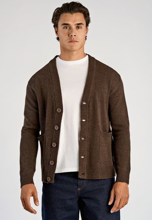 Brun uldstrik cardigan med ribbet krave og manchetter, med en række knapper, en frontlomme og en afslappet pasform over en hvid t-shirt.