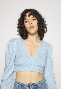 Top cropped azul com estampa floral, possui um decote em V profundo, mangas longas com efeito bufante e fecho de botão. O tecido é leve e de textura suave.
