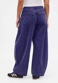 Vijolične kavbojke širokih hlačnic z visokim pasom, petimi žepi in vidnimi šivi, kombinirane s črnimi supergami z belimi črtami.