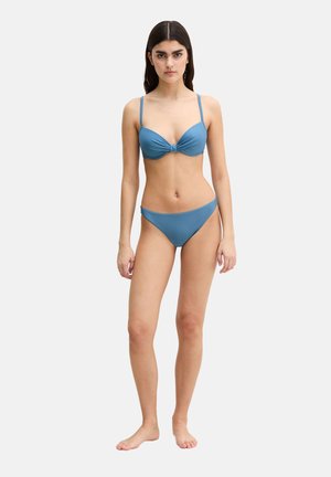Young woman standing barefoot wearing a blue bikini with thin straps against a plain light background.