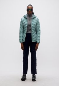 Kvinne som står, iført teal farget hettejakke med dun, grå genser med glidelås, svarte bukser, tursko og reflekterende sportssolbriller.