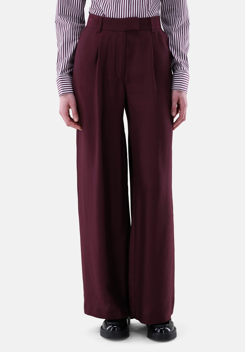 Weite Hosen aus burgunderfarbener Stoff mit Falten an der Vorderseite. Kombiniert mit einem gestreiften Hemd mit langen Ärmeln und Knopf-Manschetten.