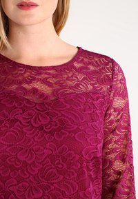 Fuchsia spetstopp med blommönster, rund halsringning och långa genomskinliga ärmar, som framhäver intrikata designdetaljer och texturerad tyg.