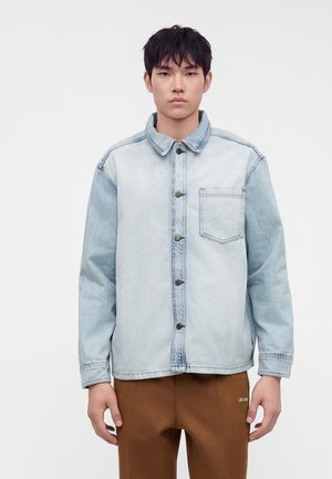 Junger Mann trägt ein hellblaues Jeanshemd mit Brusttasche und brauner Hose, steht vor einem schlichten weißen Hintergrund.
