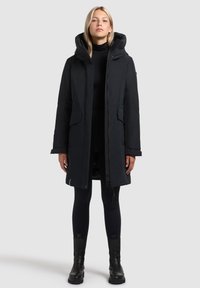 Parka neagră cu glugă, fermoar frontal și buzunare cu clapetă. Oferă o croială lejeră și un material texturat. Purtați-o peste un top negru și colanți.