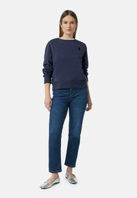 Marineblauwe sweatshirt met ronde hals, ribgebreide manchetten en een klein geborduurd logo op de borst, gecombineerd met recht gesneden denim jeans en zilveren ballerina's.
