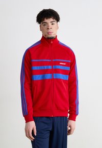 Giacca rossa con zip e strisce blu. Realizzata in tessuto liscio, presenta un colletto alto e un design a tre strisce sulle maniche. Logo Adidas visibile.