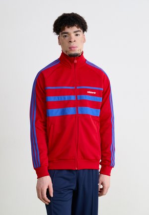 Chaqueta roja con cremallera y rayas azules. Confeccionada en tejido suave, presenta un cuello alto y un diseño de tres rayas en las mangas. Logo de Adidas exhibido.