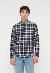 Καρό πουκάμισο σε ναυτικό μπλε και λευκό με μακριά μανίκια, γιακά με κουμπιά, και εμφανή χρυσά κουμπιά κατά μήκος του μπροστινού πλακέ.