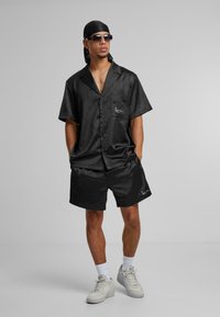 Sort satin kortærmet skjorte med lomme og logo, matchende sorte shorts, båret med hvide sneakers og solbriller, sat mod en grå baggrund.