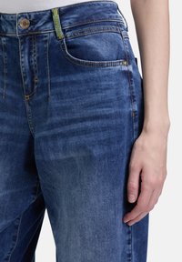 Jeans aus Denim in einem mittleren Blauton, mit einem Knopfverschluss, einer Fronttasche und grünen Nähdetails am Bund.