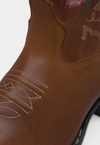 Καφέ δερμάτινη καουμπόικη μπότες με διακοσμητικό λευκό ραφτό στο δάχτυλο και πολύχρωμο κεντητό λουλουδάτο σχέδιο κοντά στον αστράγαλο.