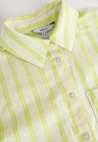 Camicia beige chiaro con righe verdi, colletto, bottoni e tasca frontale. Il materiale appare strutturato con una finitura morbida. Etichetta del marchio visibile all'interno.