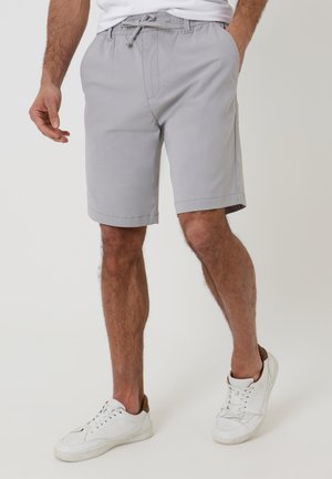 Mand iført lysegrå shorts med snøre og hvide sneakers, stående med afslappede hænder, neutral baggrund.