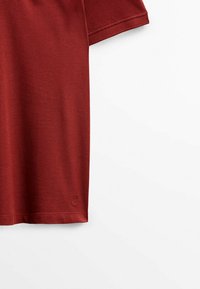 Polo manches courtes rouge rouille en tissu texturé avec petit logo brodé près de l'ourlet inférieur sur fond blanc.
