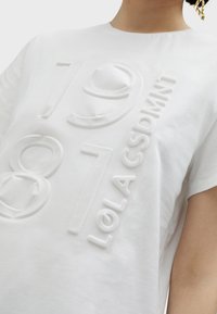 Camiseta de algodón blanca con diseño de texto en relieve y en relieve que presenta los números "1981" y la palabra "LOLACSDMNT". Mangas cortas y escote redondo.