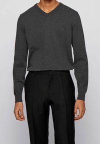 Dunkelgrauer V-Ausschnitt-Pullover aus weichem Stoff, mit langen Ärmeln und einem dezenten Logo auf der Brust, kombiniert mit schwarzer Hose.