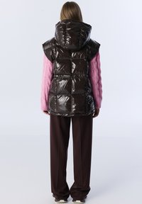Brązowa, błyszcząca kamizelka puchowa z kapturem i bez rękawów, w połączeniu z różowym swetrem w warkocze i brązowymi spodniami.