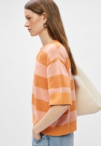 Gestreepte gebreide trui in oranje en lichtroze, met korte mouwen en een ronde halslijn. Model draagt een lichte tas over haar schouder.