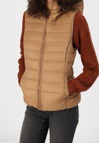 Veste matelassée beige avec fermeture éclair, dotée d'une capuche et de manches côtelées orange. Texture de surface avec des coutures horizontales.